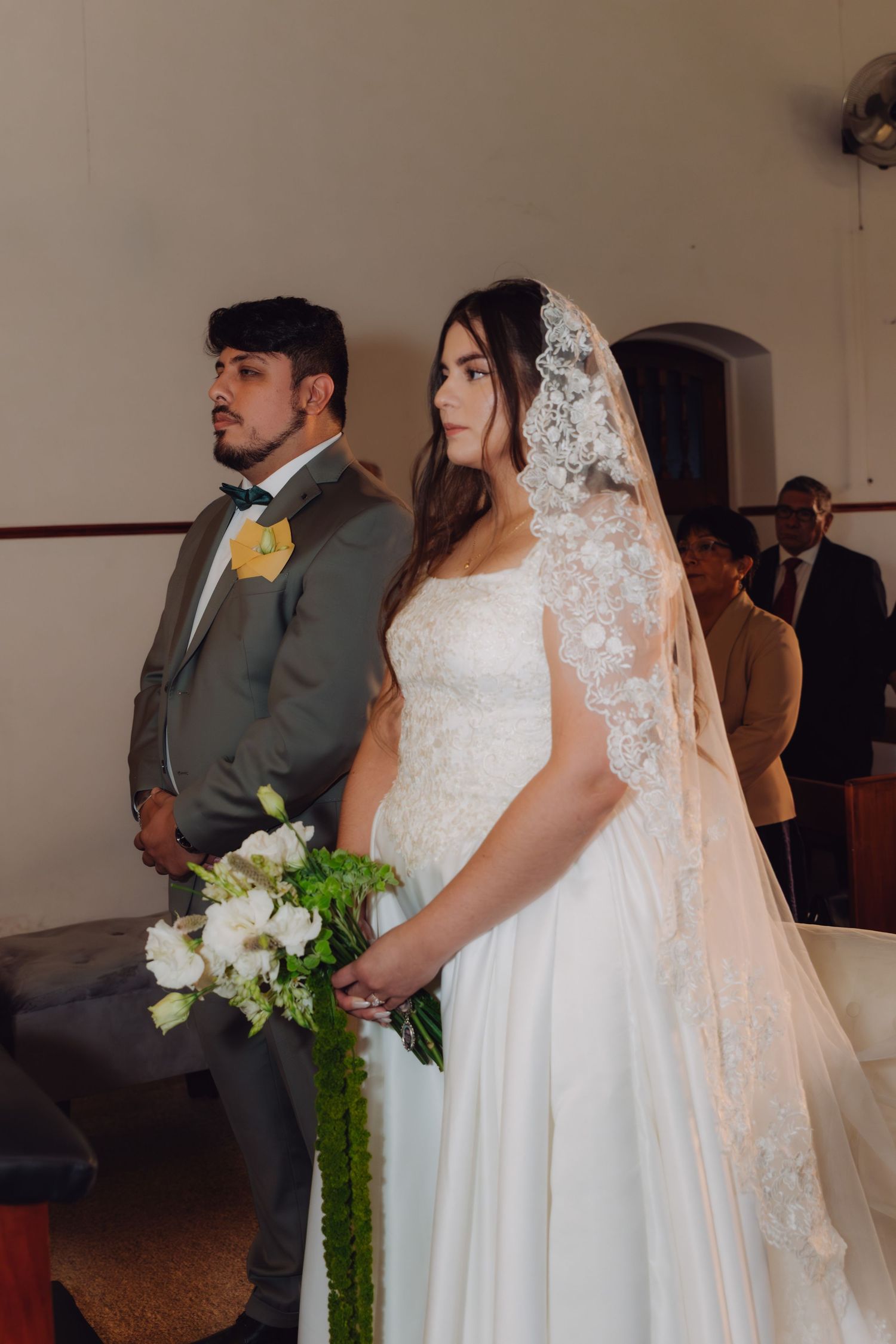 Bride in white ball gown with lace veil and groom in gray suit stand at altar during church ceremony.