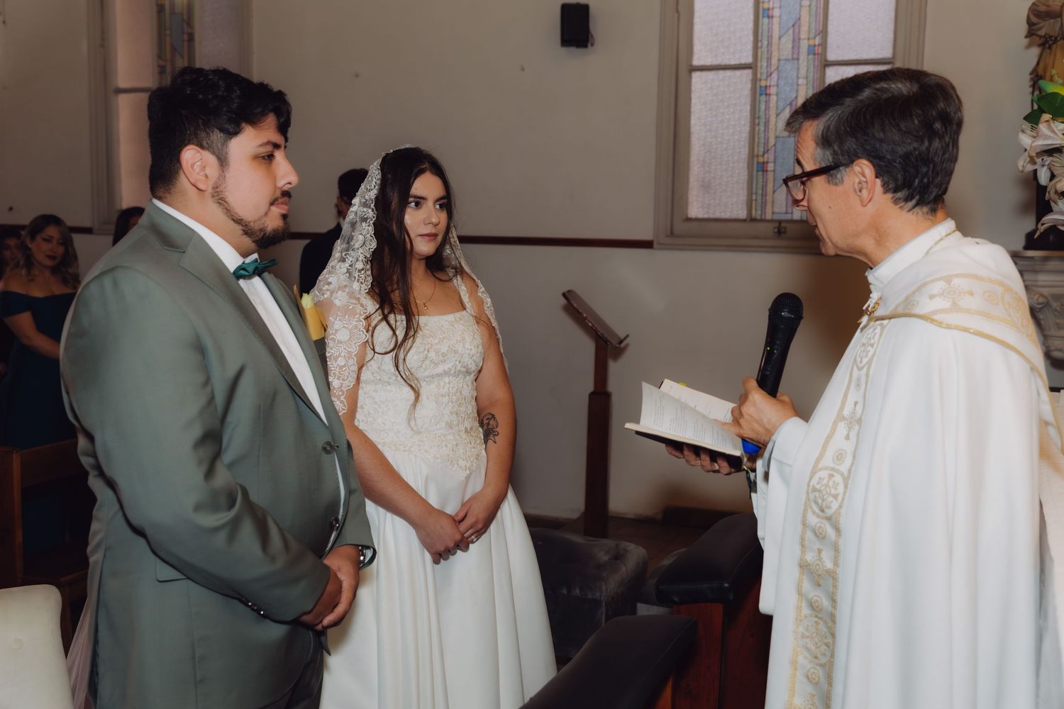 Priest in white vestments reads from book to bride and groom during Catholic wedding ceremony.