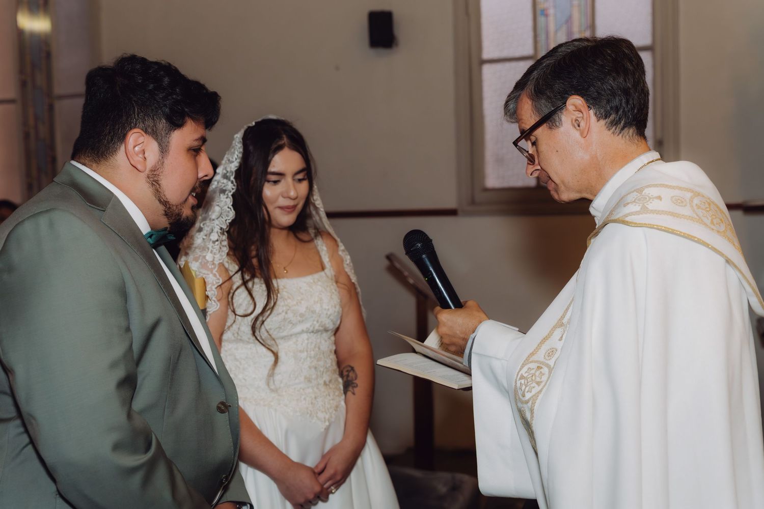 Officiant reads vows to bride in lace veil and groom in gray suit at church wedding altar.