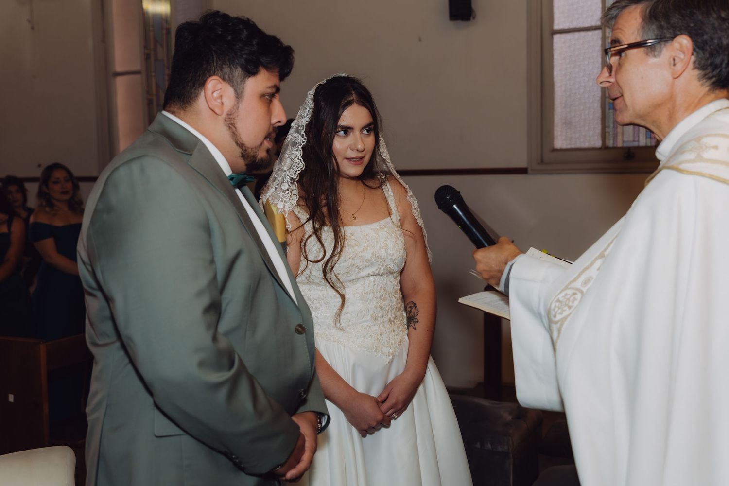 Bride and groom bow heads as priest holds microphone during Catholic wedding ceremony in church.