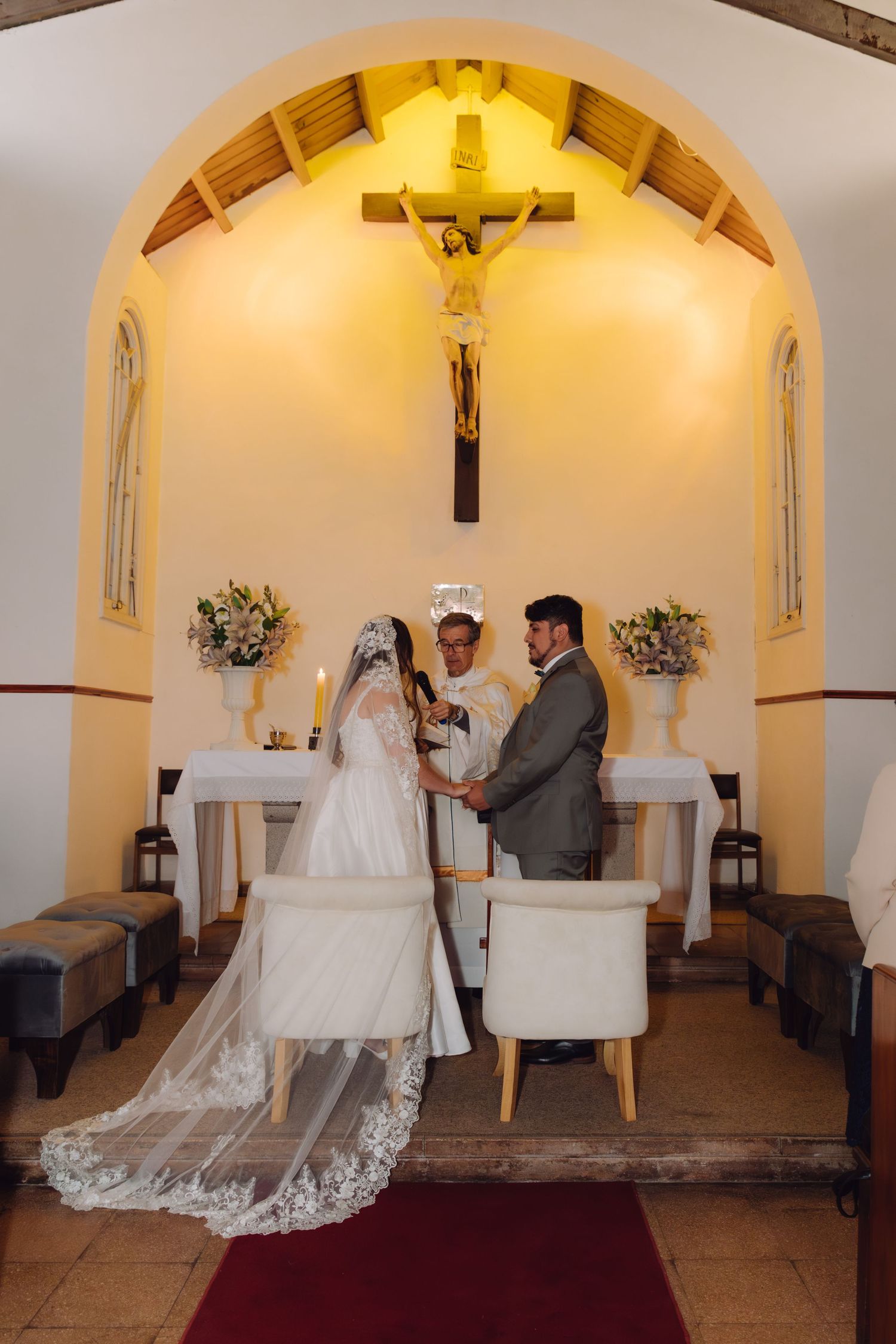 Bride and groom hold hands at altar beneath crucifix during Catholic church wedding ceremony.