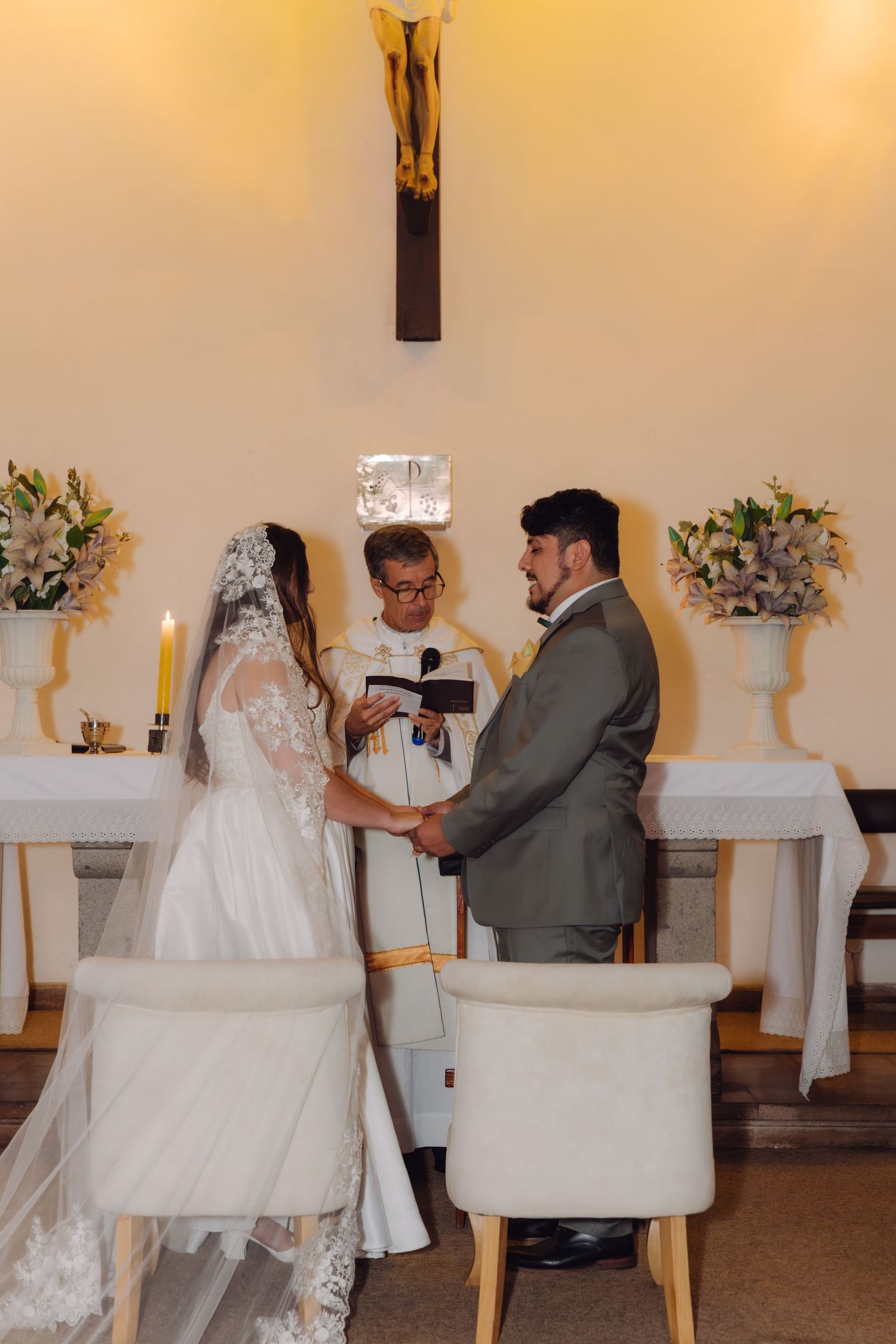Bride and groom exchange vows holding hands before priest at decorated church altar with candles.
