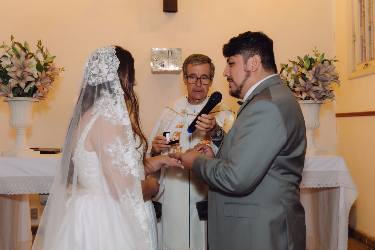 Groom places wedding ring on bride's finger during church ceremony with priest.