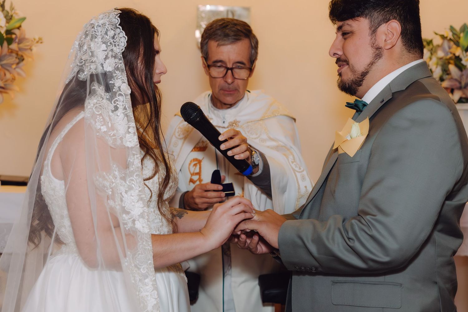 Bride slides wedding ring onto groom's finger as priest holds microphone.