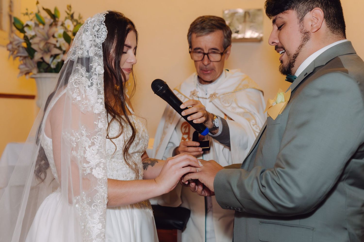 Priest holds microphone as bride and groom complete ring exchange ceremony.