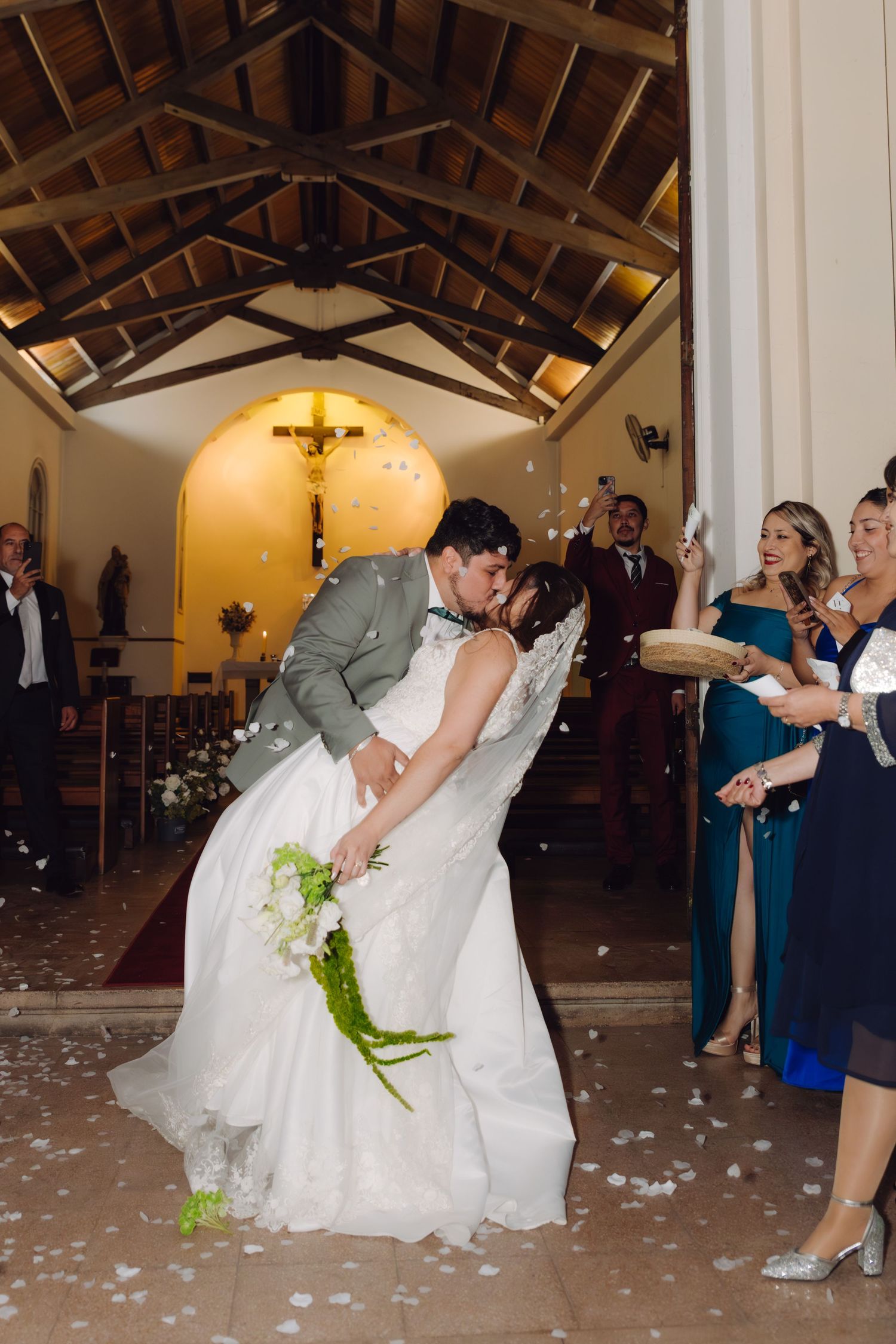 Groom dips his bride for a romantic kiss as guests toss white flower petals at their church exit.