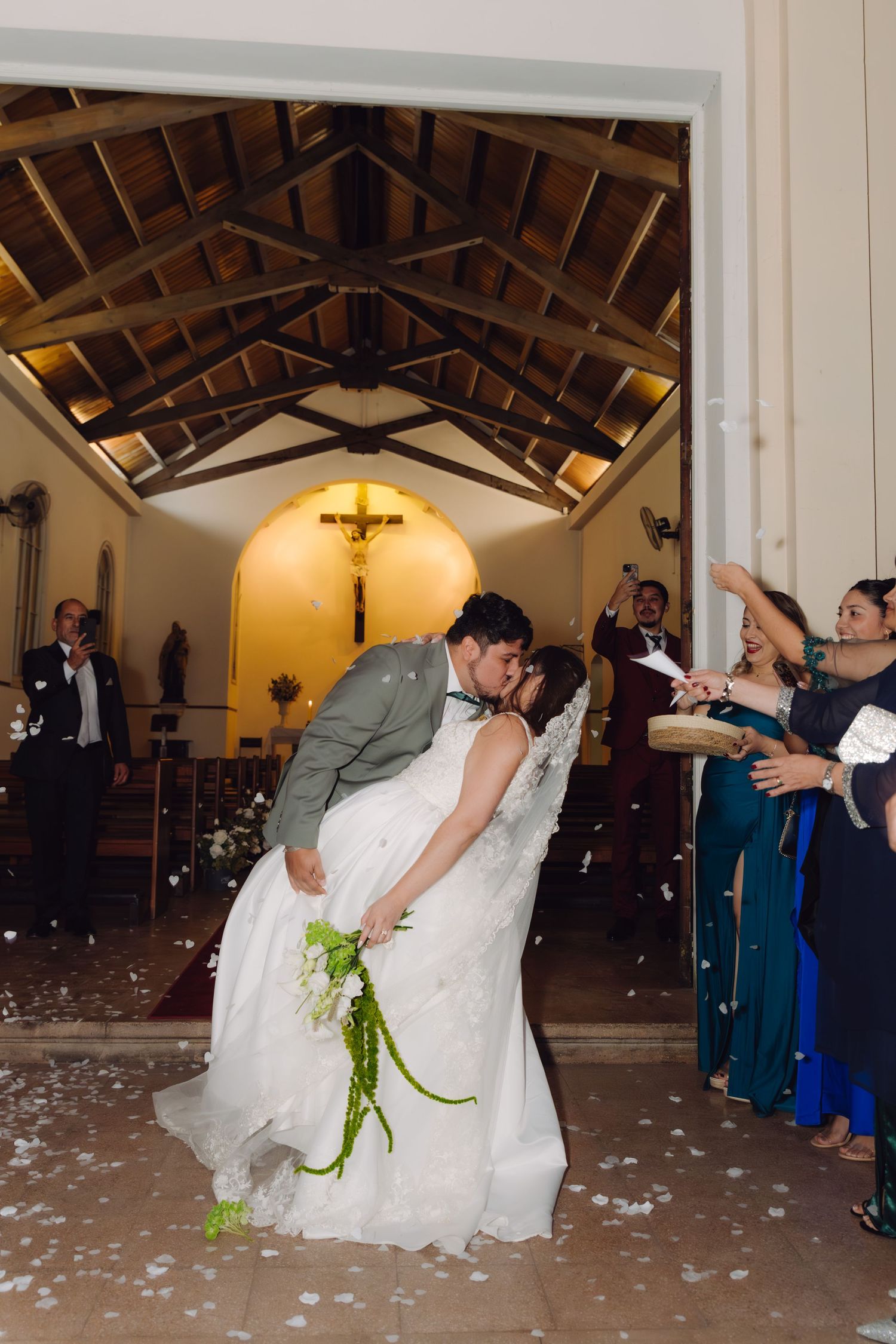 Newlyweds share a passionate dip kiss at church entrance as guests throw white petals in celebration.