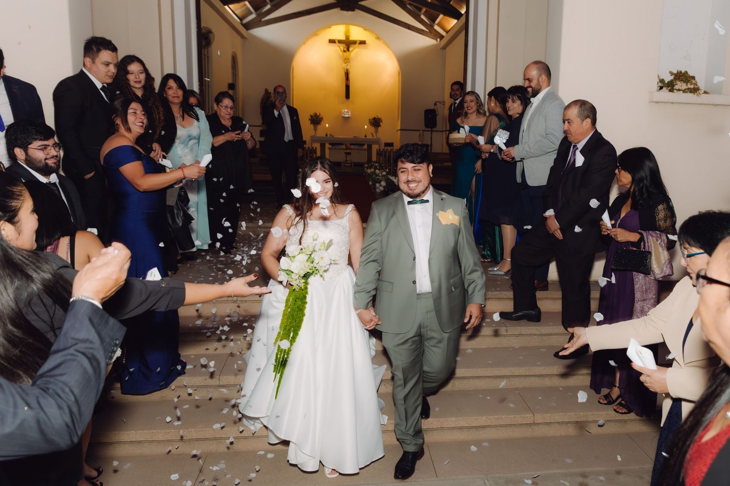Bride and groom descend church steps as smiling guests line up to shower them with flower petals.