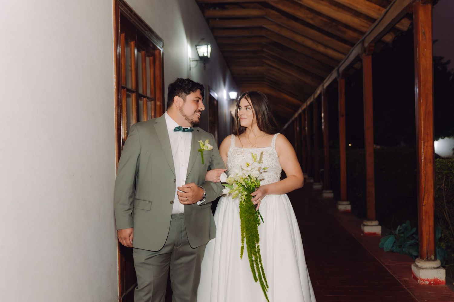 Elegant bride and groom walk together along a covered outdoor corridor after their evening wedding.