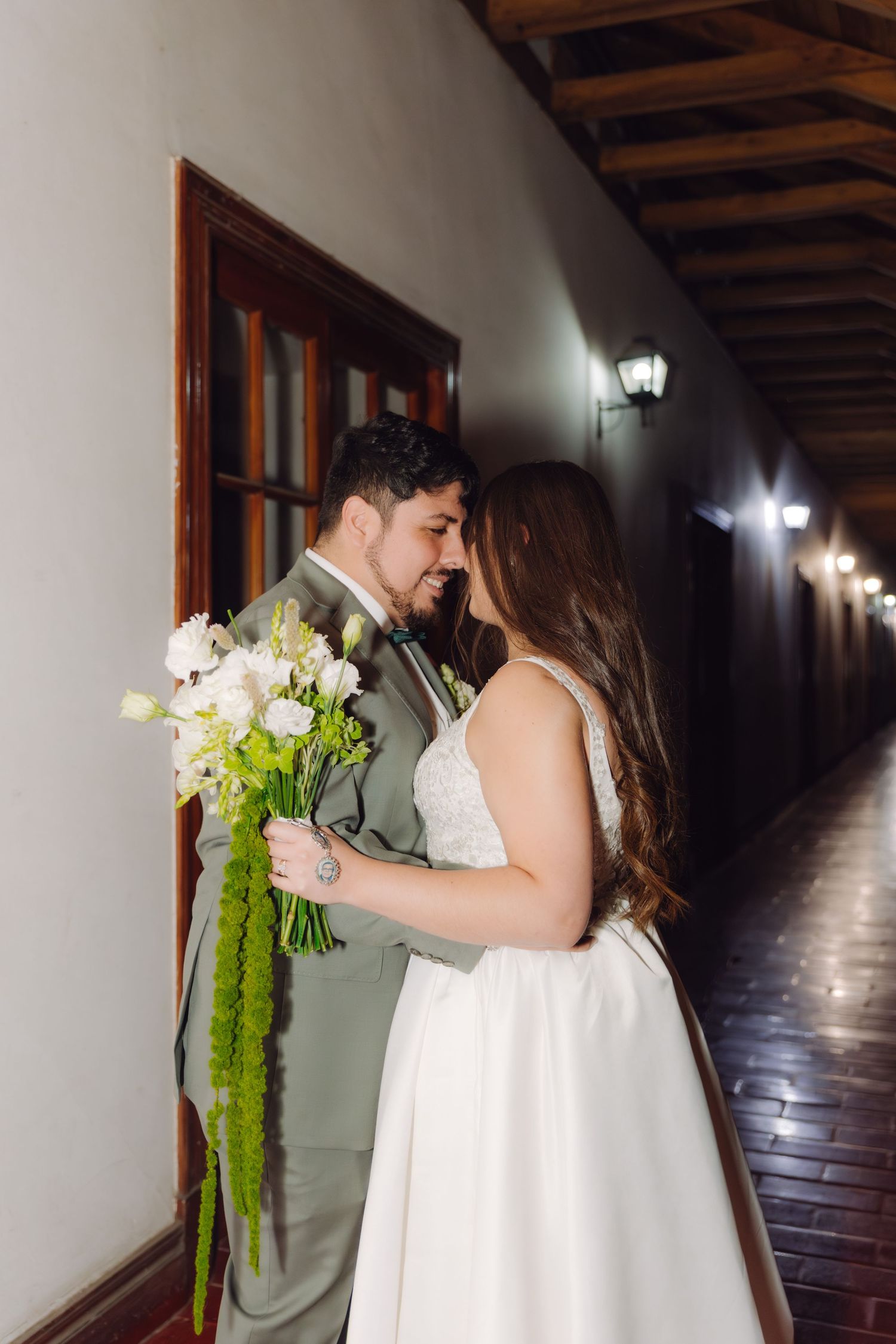 Bride and groom share an intimate moment leaning close together in a warmly lit outdoor corridor.
