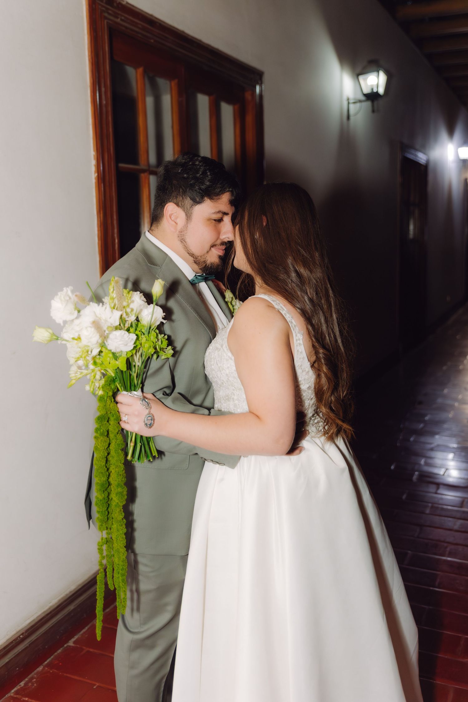 Bride and groom share an intimate moment in a hallway, with the groom holding a white and green wedding bouquet.