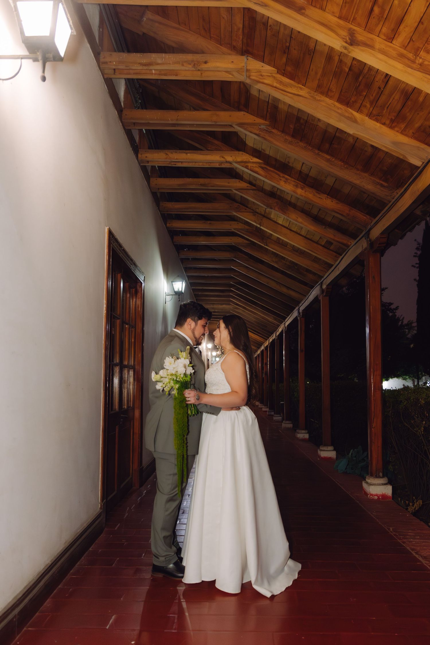 Newlyweds stand close together under a wooden beamed corridor at night, bride holding a white floral bouquet.