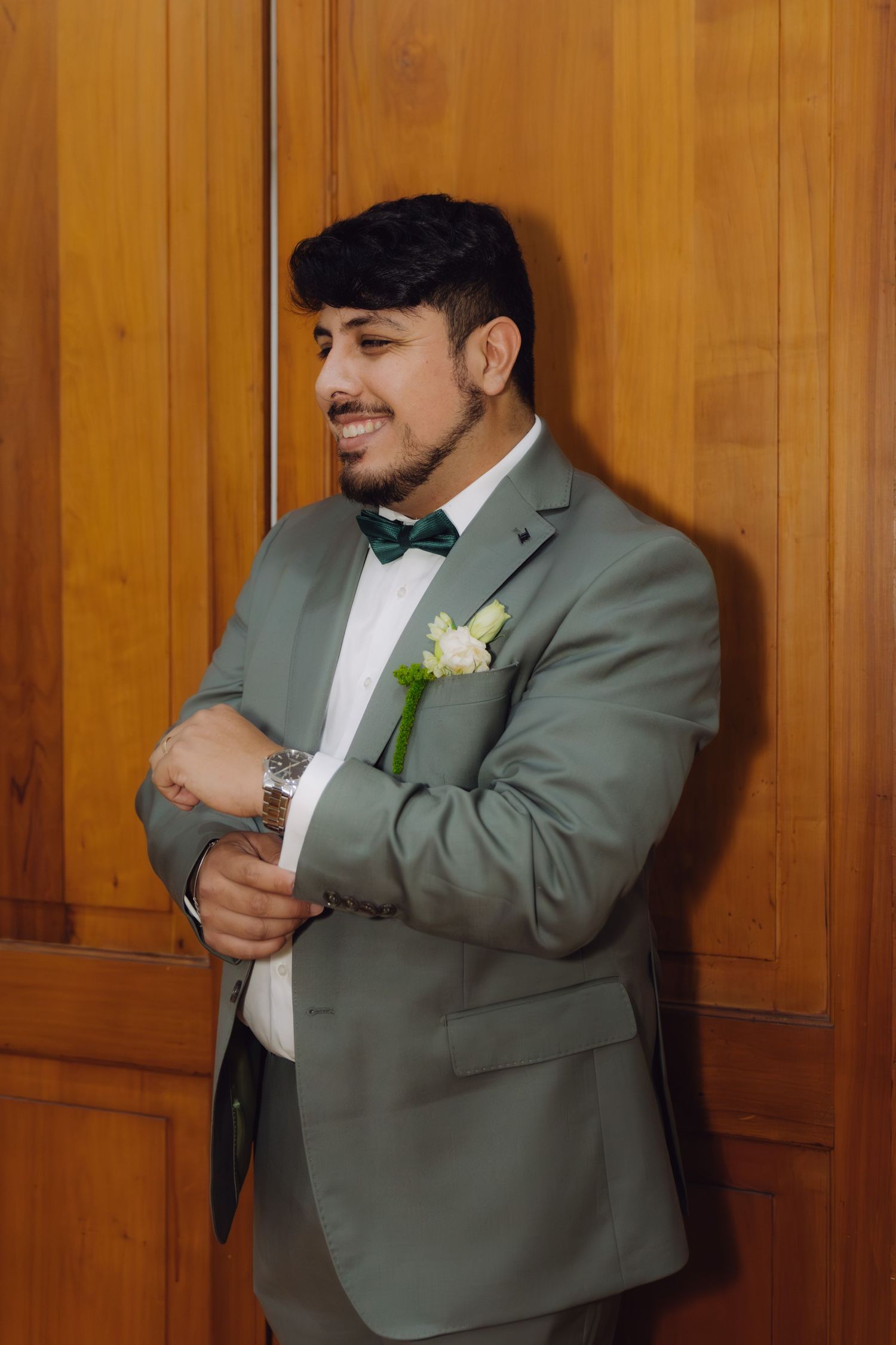 Smiling groom in a sage green suit with green bow tie and white boutonniere adjusts his cufflinks indoors.