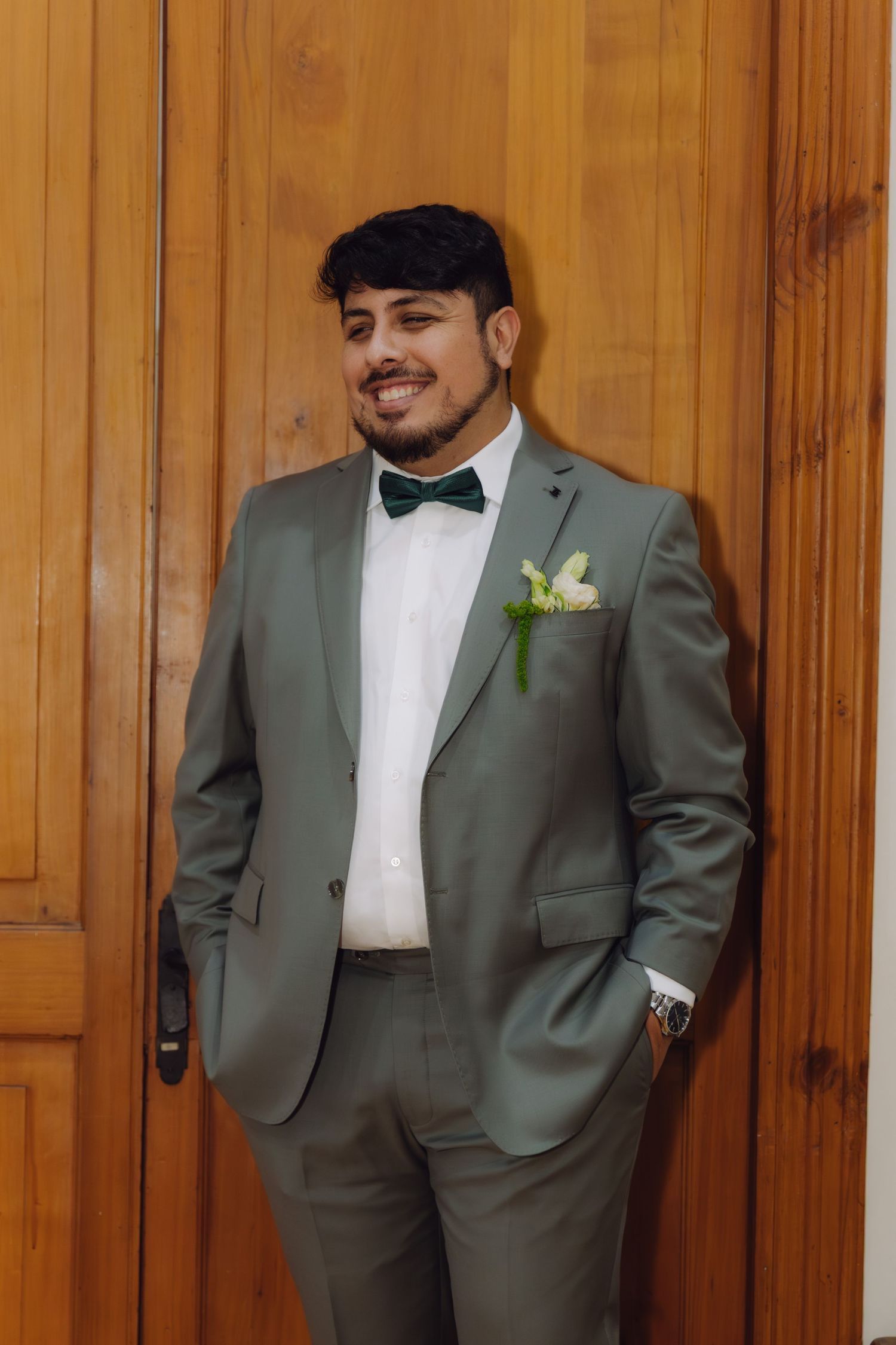 Handsome groom in sage green suit and dark green bow tie poses confidently against wooden doors.