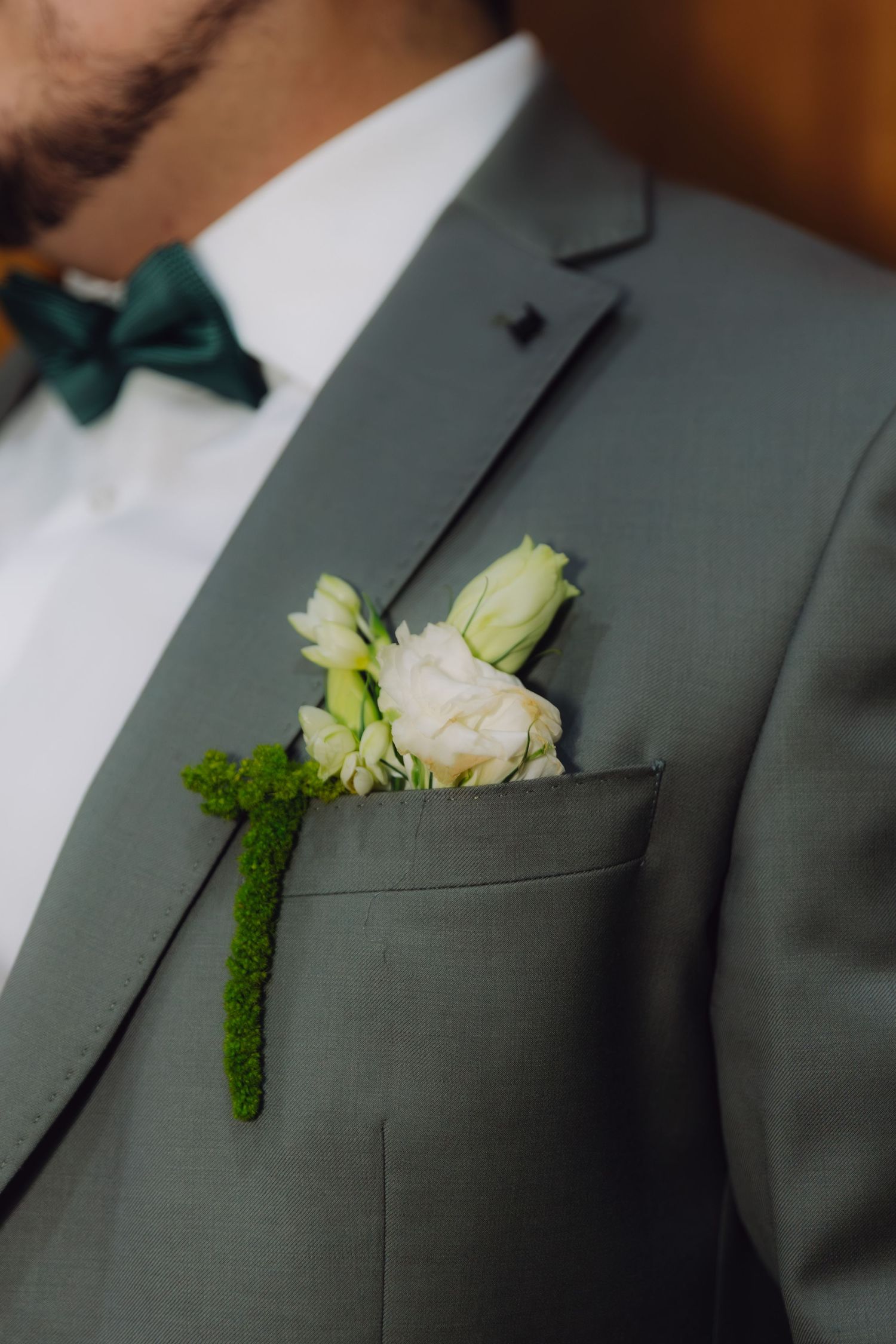 Close-up of a groom's sage green suit lapel featuring a white rose and green moss boutonniere.