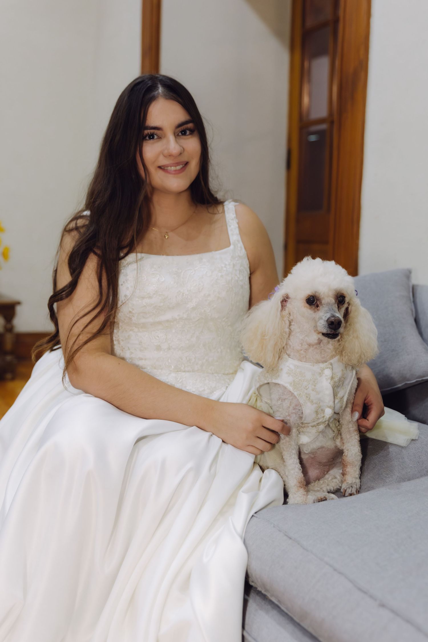 Smiling bride in white lace wedding gown sits on a sofa with her white poodle dressed in a bow.