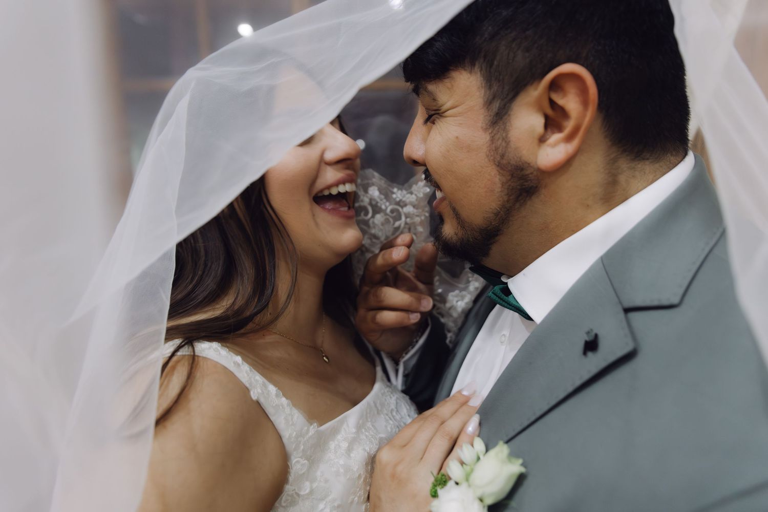 Bride and groom share a joyful moment under a wedding veil with a small cat between them.