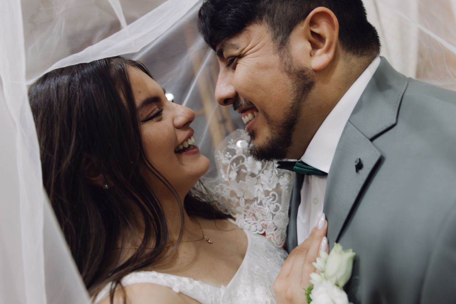 Happy bride and groom smile at each other closely under a flowing white wedding veil.