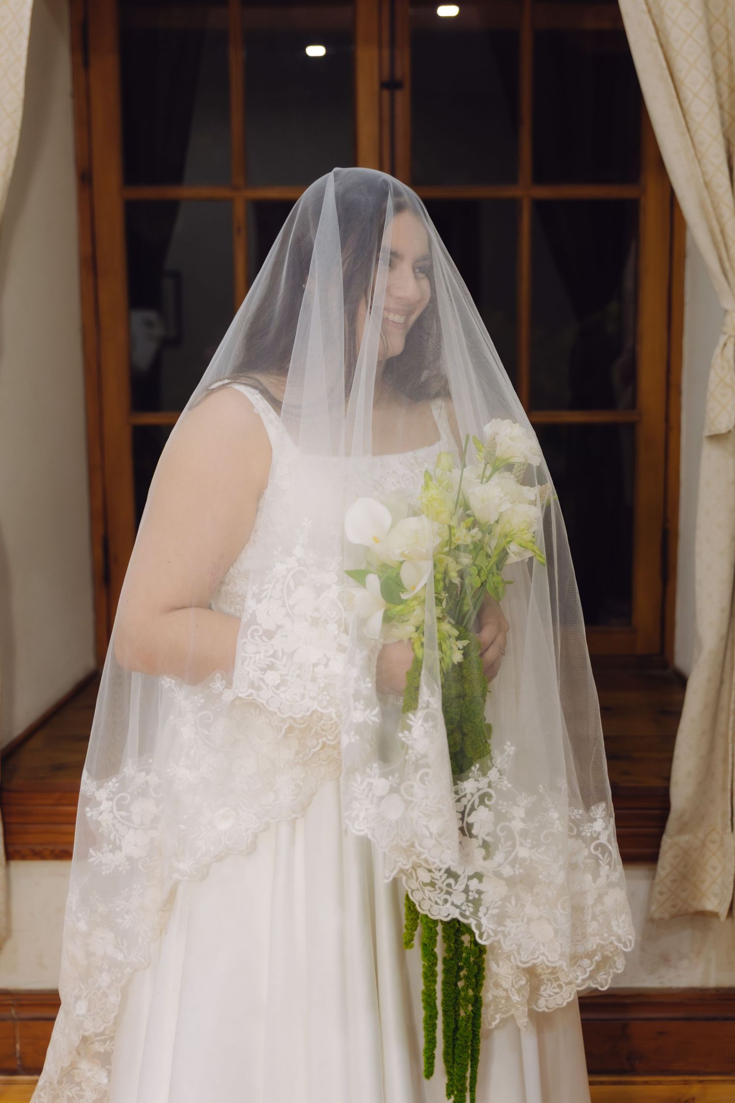 Smiling bride in white lace gown holds a bouquet of white flowers under her veil.