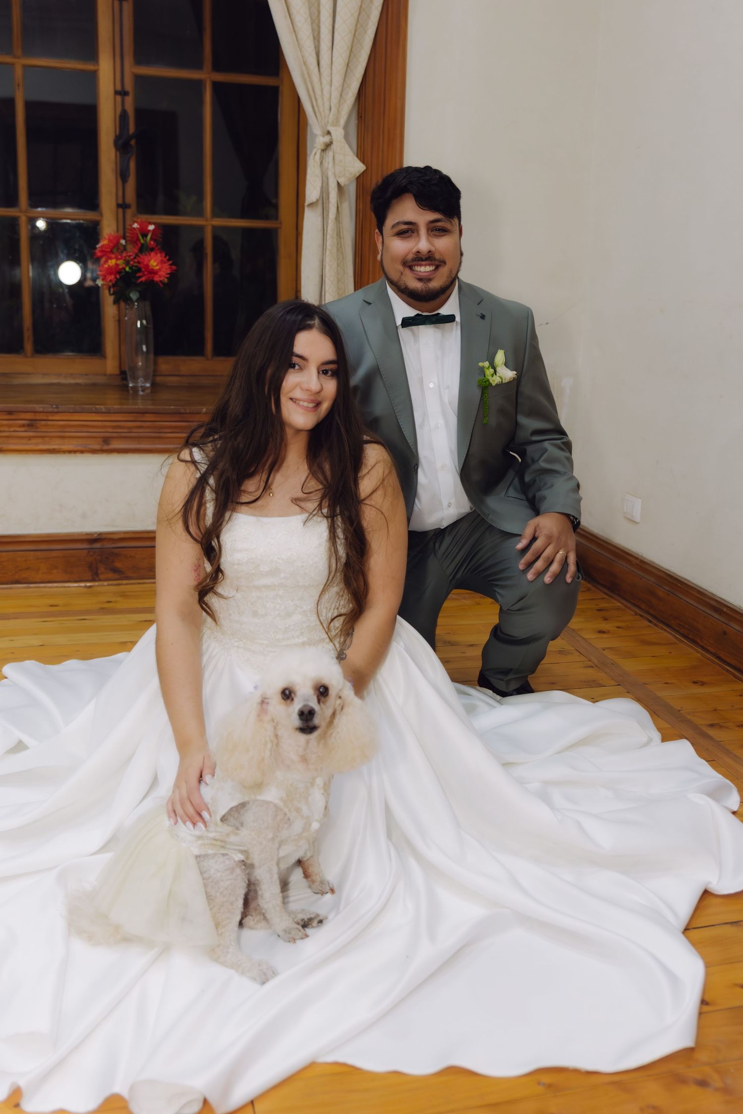 Bride and groom pose with their white poodle on a wooden floor at their wedding.
