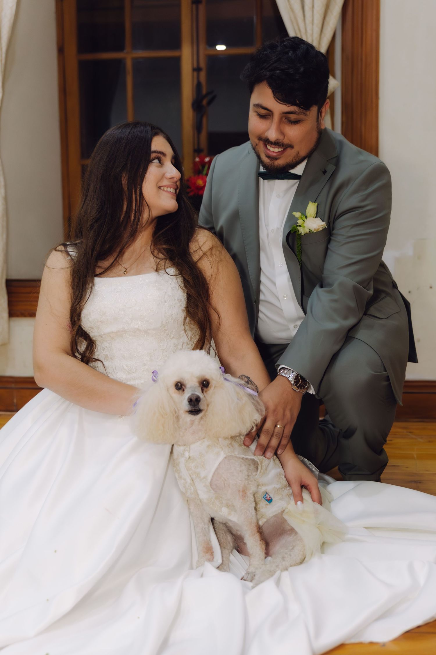 Newlyweds sit together on the floor smiling at their white poodle in wedding attire.