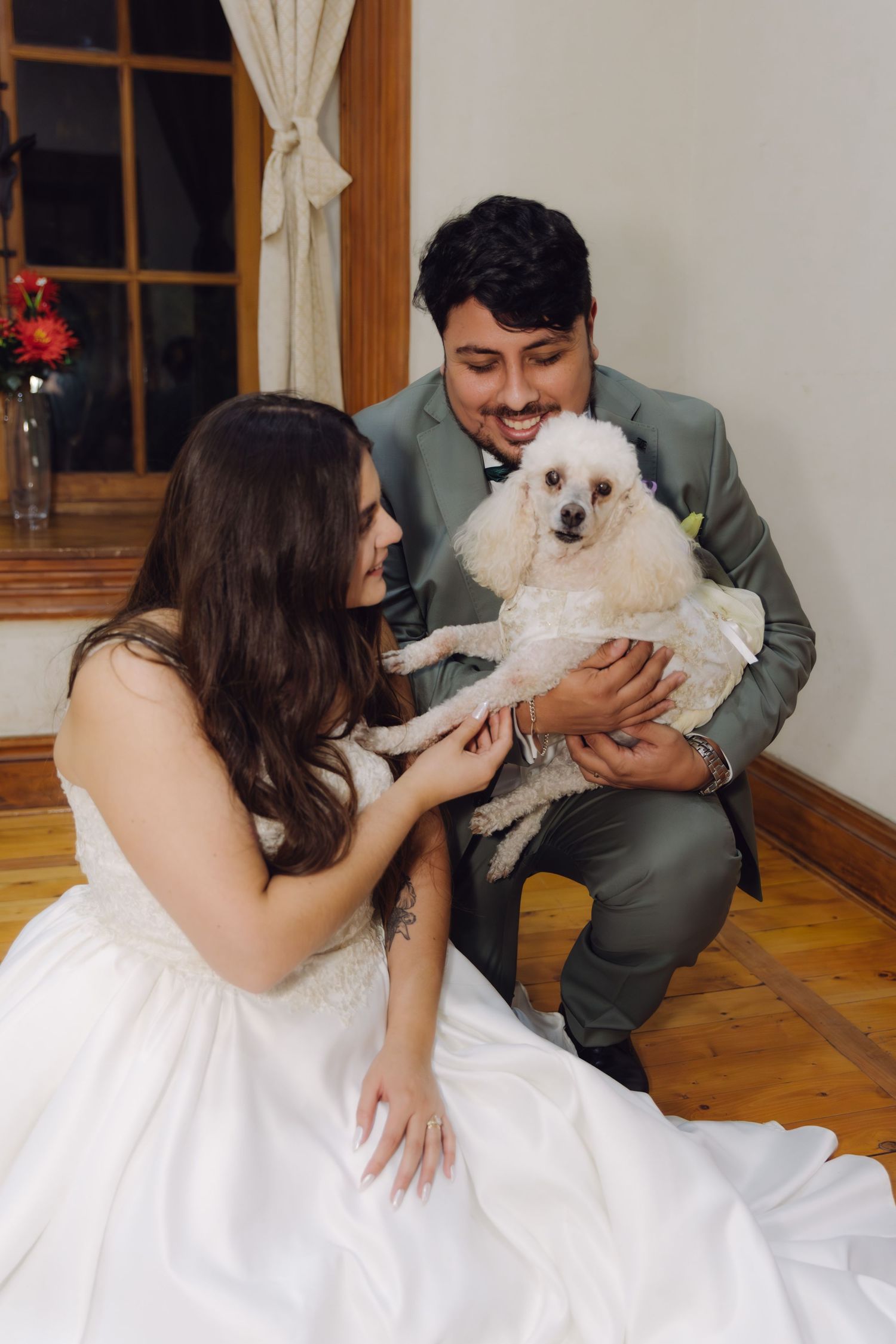 Bride and groom hold their white poodle together while sitting on the floor indoors.