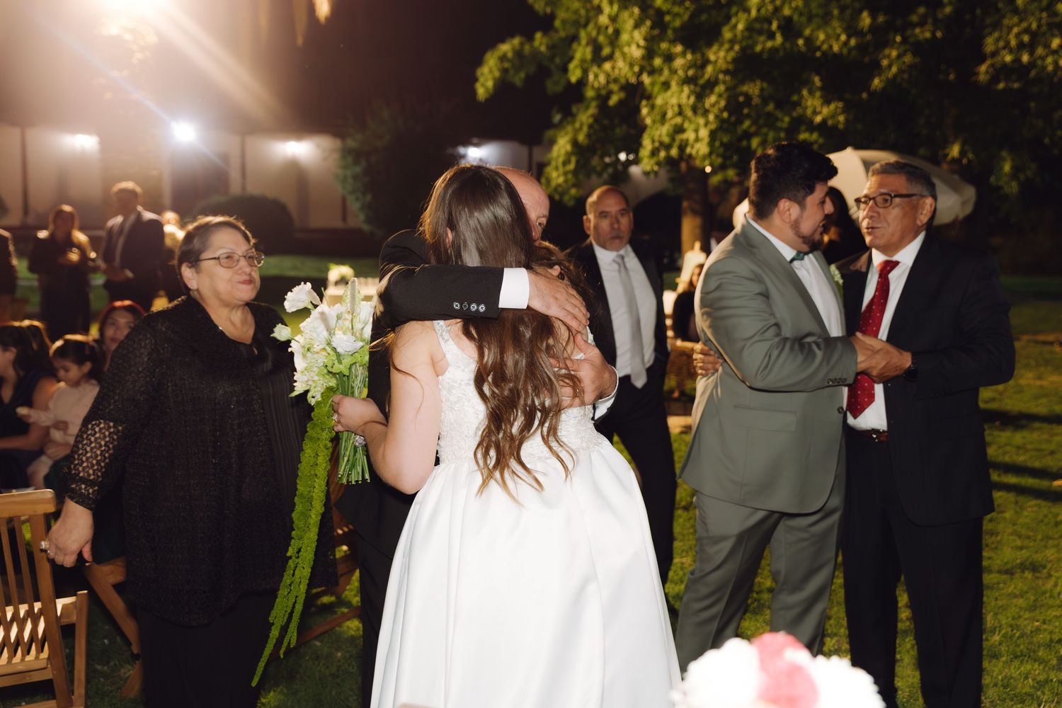 Wedding guests embrace the bride and groom during an outdoor evening reception ceremony.