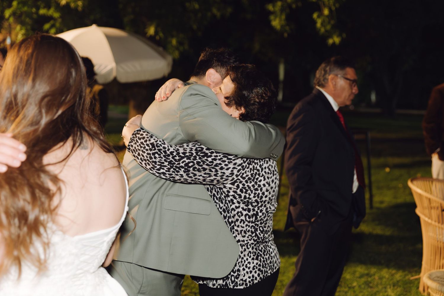 Groom in sage suit shares a warm hug with a guest at an outdoor wedding reception.