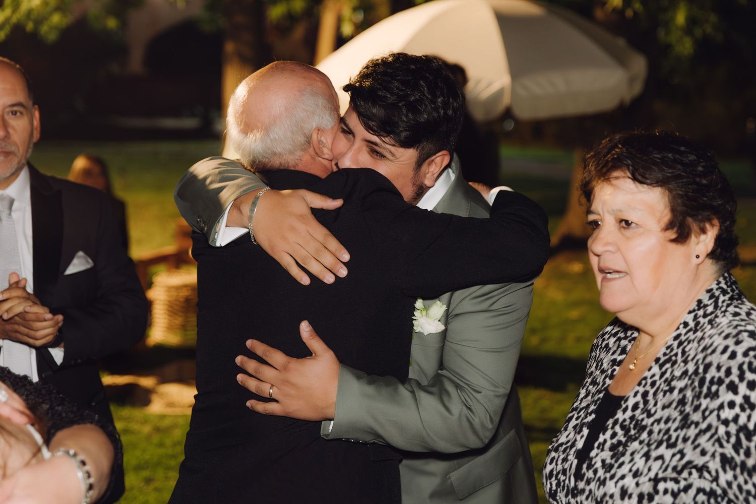 Groom in gray suit shares emotional hug with elderly guest at outdoor wedding reception at night.