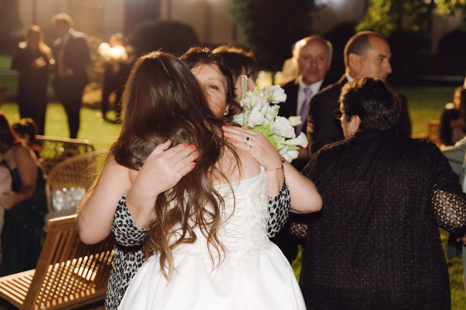 Bride in white gown holding white flowers receives tender hug from guest at nighttime wedding.