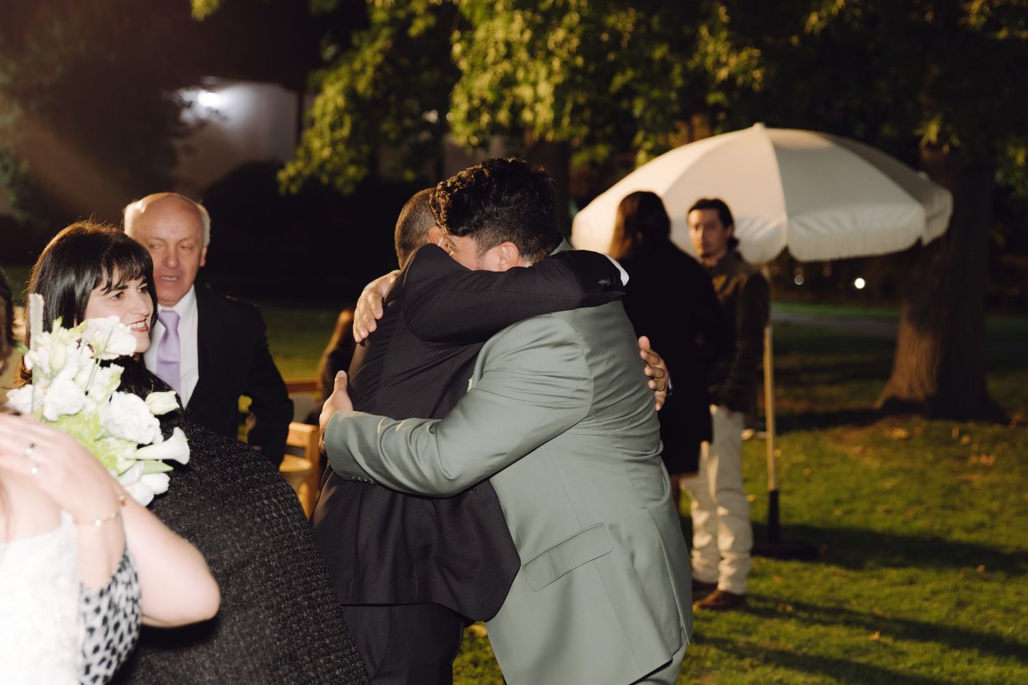 Wedding guests embrace warmly during outdoor evening reception with white umbrella in background.