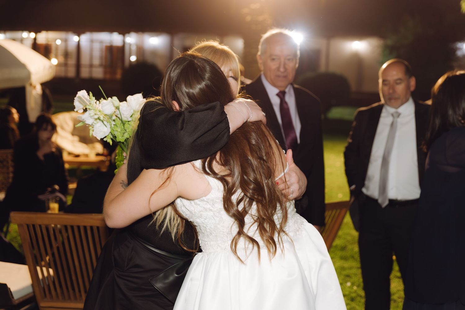 Bride holding white bouquet shares heartfelt embrace with guest while others look on at evening wedding.