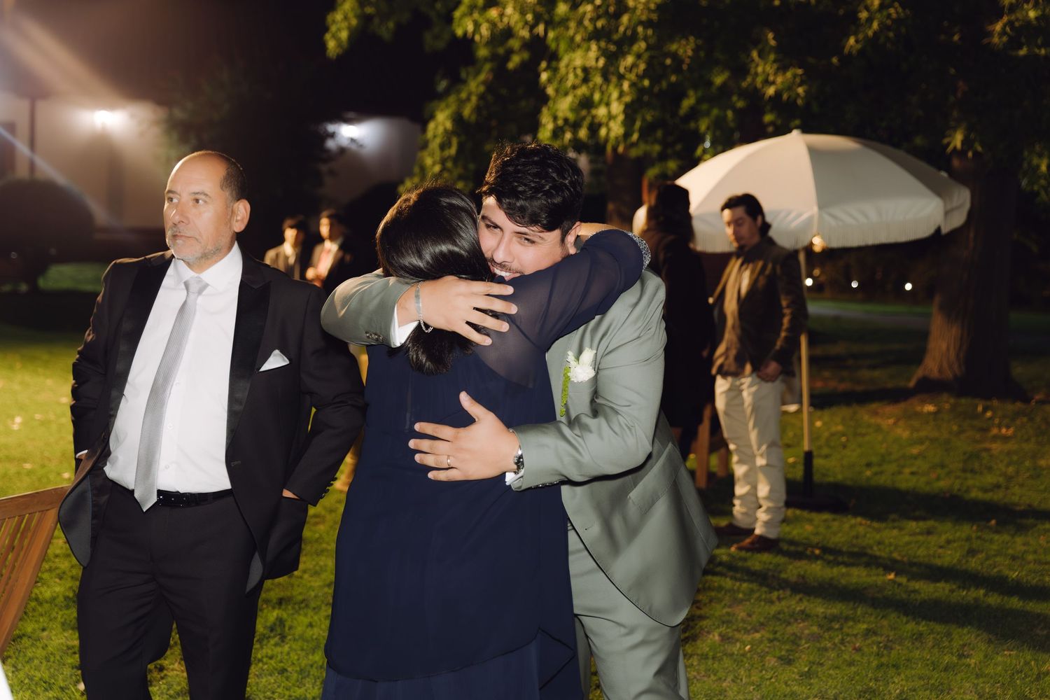 Groom in gray suit embraces female guest warmly at elegant outdoor evening wedding reception.
