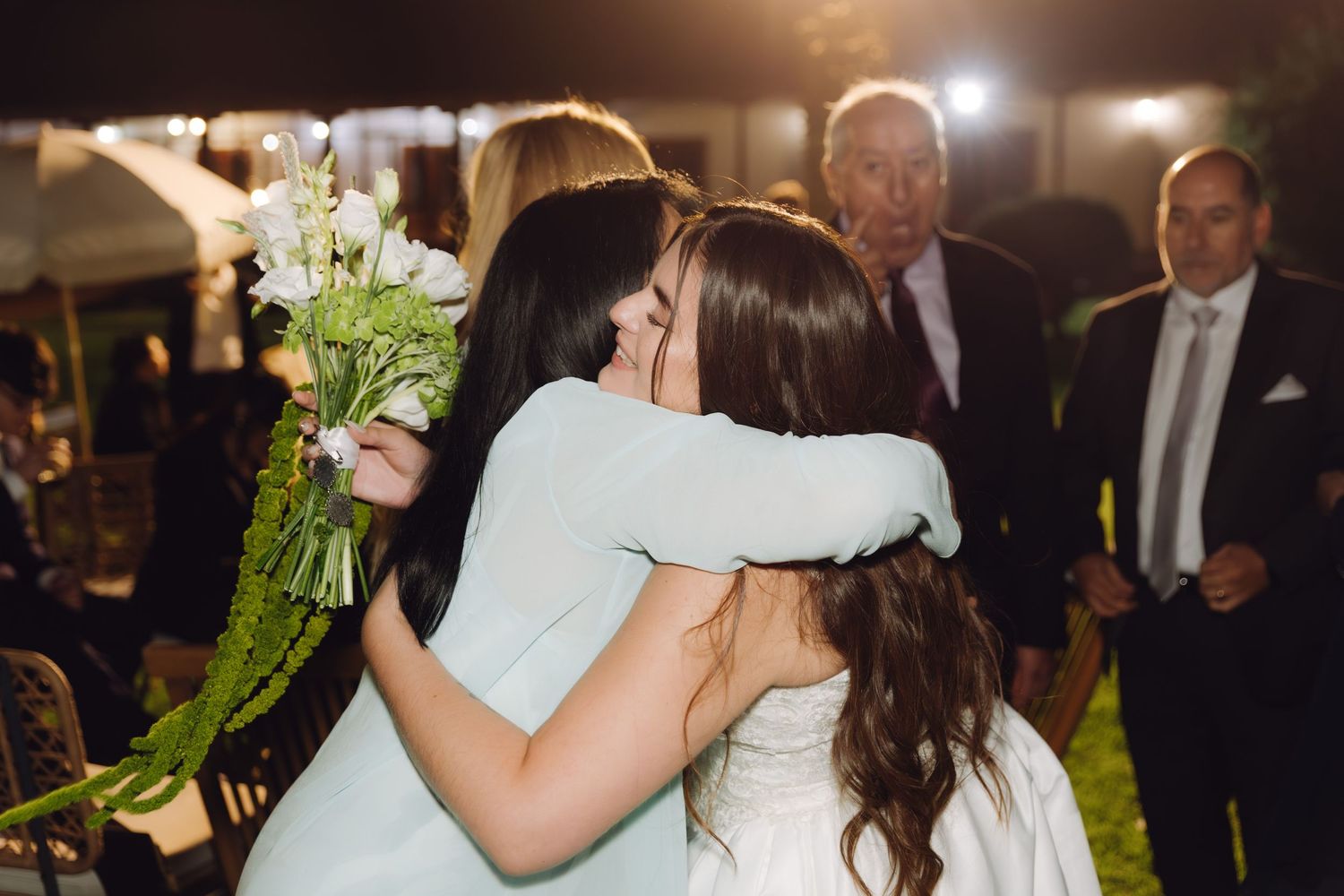 Bride and bridesmaid in mint dress share emotional hug holding white flowers at outdoor night wedding.