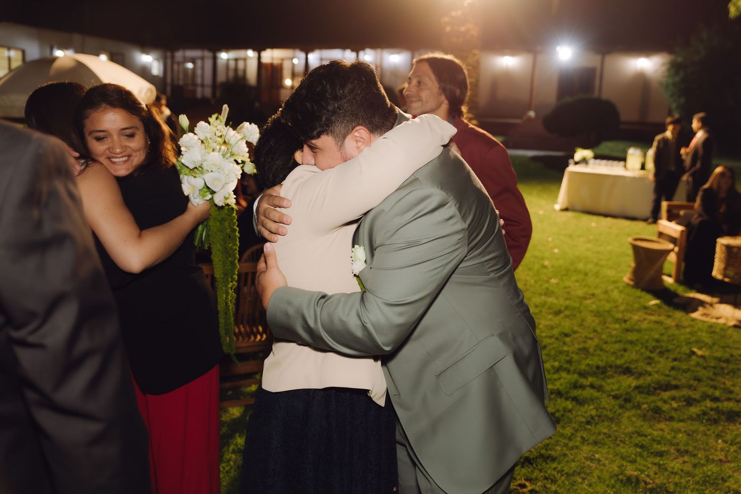 Groom hugs wedding guest warmly while smiling woman holds white bouquet at nighttime outdoor reception.