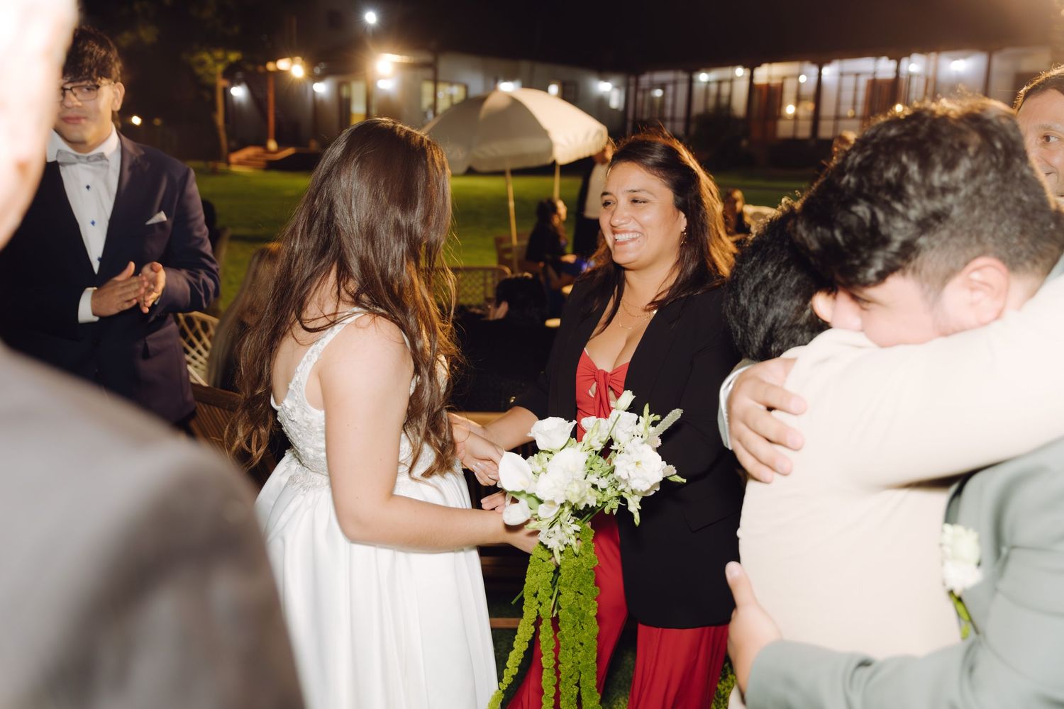 Bride in white dress receives bouquet while guests embrace at outdoor evening wedding celebration.