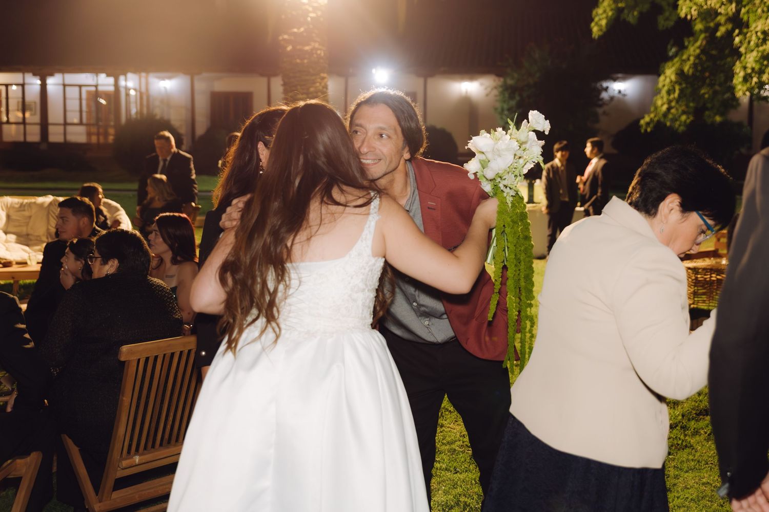 Bride in white gown receives hug from guest in green outfit at outdoor evening wedding reception.