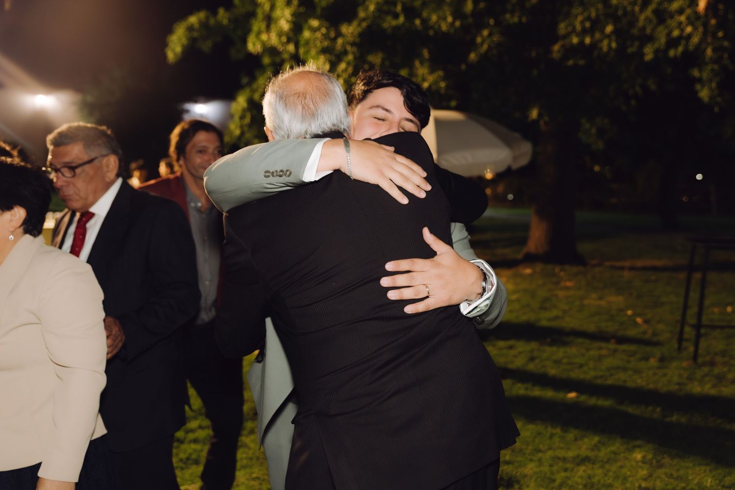 Groom in gray suit shares warm embrace with older gentleman at outdoor nighttime wedding reception.