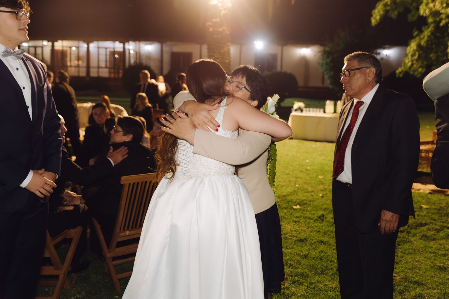 Bride in white gown receives warm hug from guest at outdoor evening wedding reception.