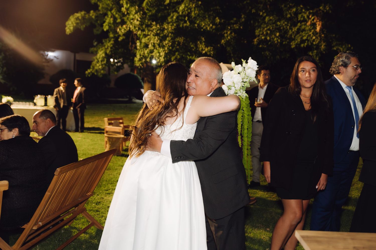 Bride in white dress shares heartfelt hug with older gentleman at outdoor wedding reception.
