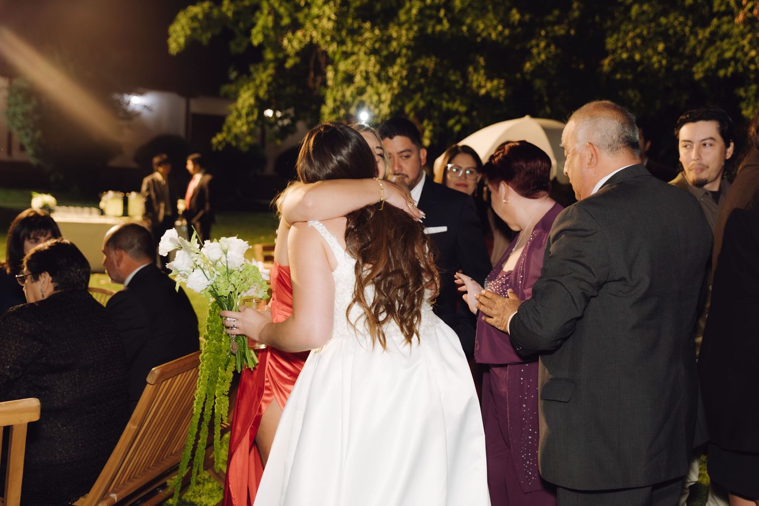 Bride holding white floral bouquet receives congratulatory hug from guest at outdoor wedding.