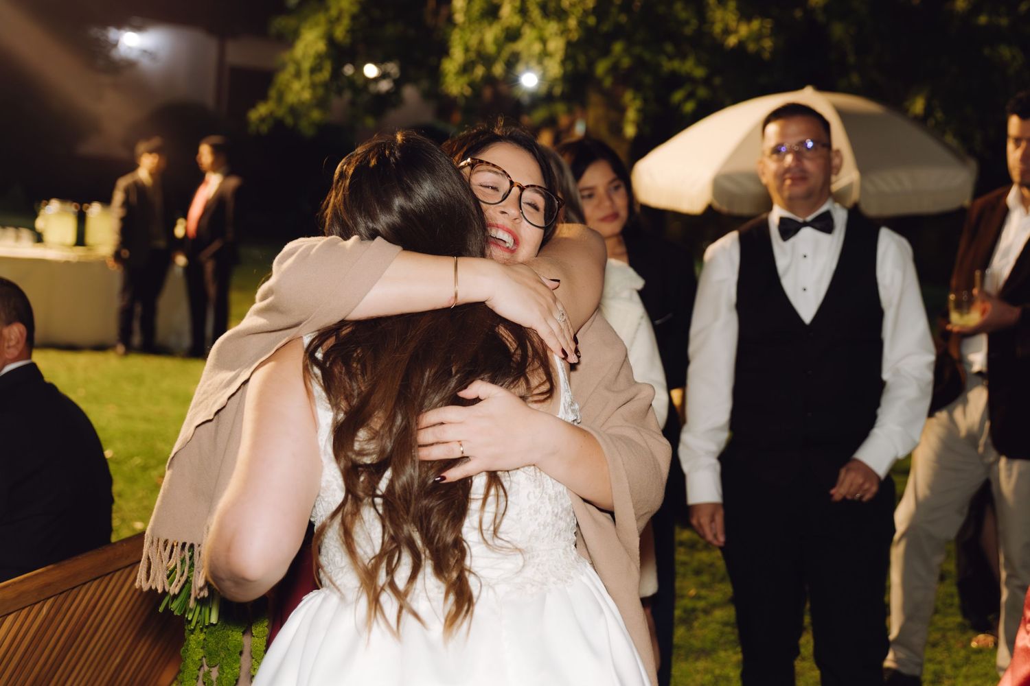 Bride in white gown receives warm group hug from emotional guests at outdoor wedding reception.