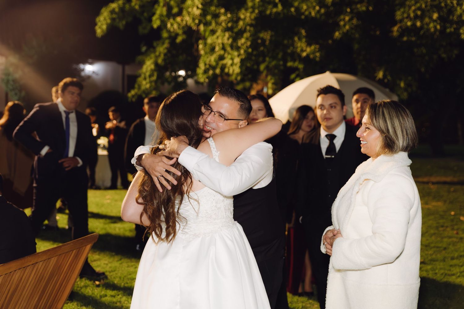Bride hugs smiling guest warmly while others look on at outdoor nighttime wedding reception.