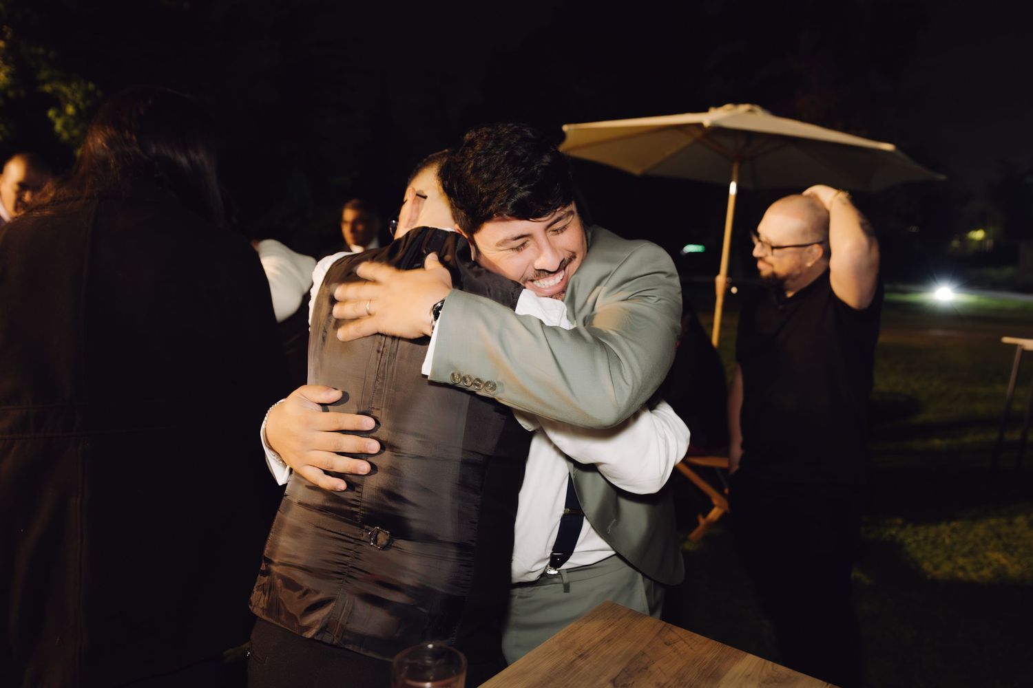 Two men in suits share a joyful embrace at an outdoor evening wedding reception.
