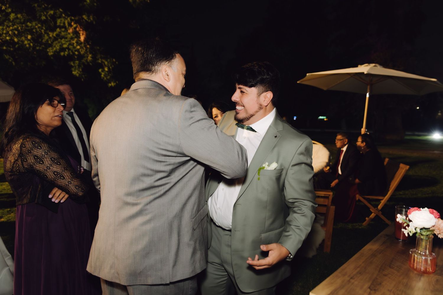 Two men in gray suits greet each other warmly at an outdoor nighttime wedding reception.