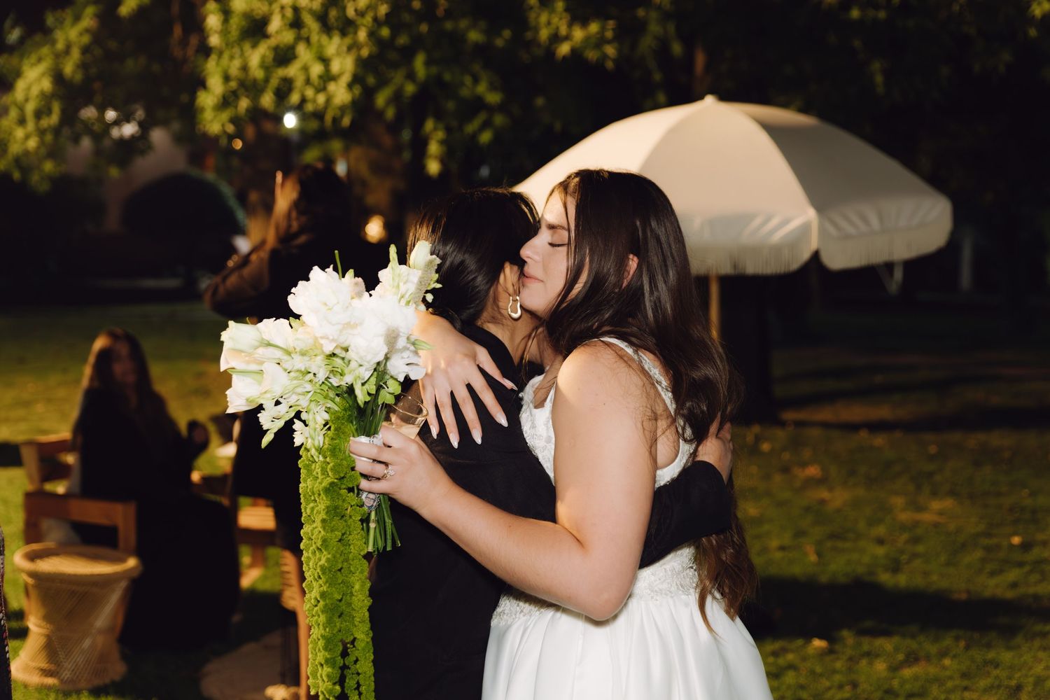Bride holding tall white floral arrangement shares tender kiss with guest at outdoor wedding.