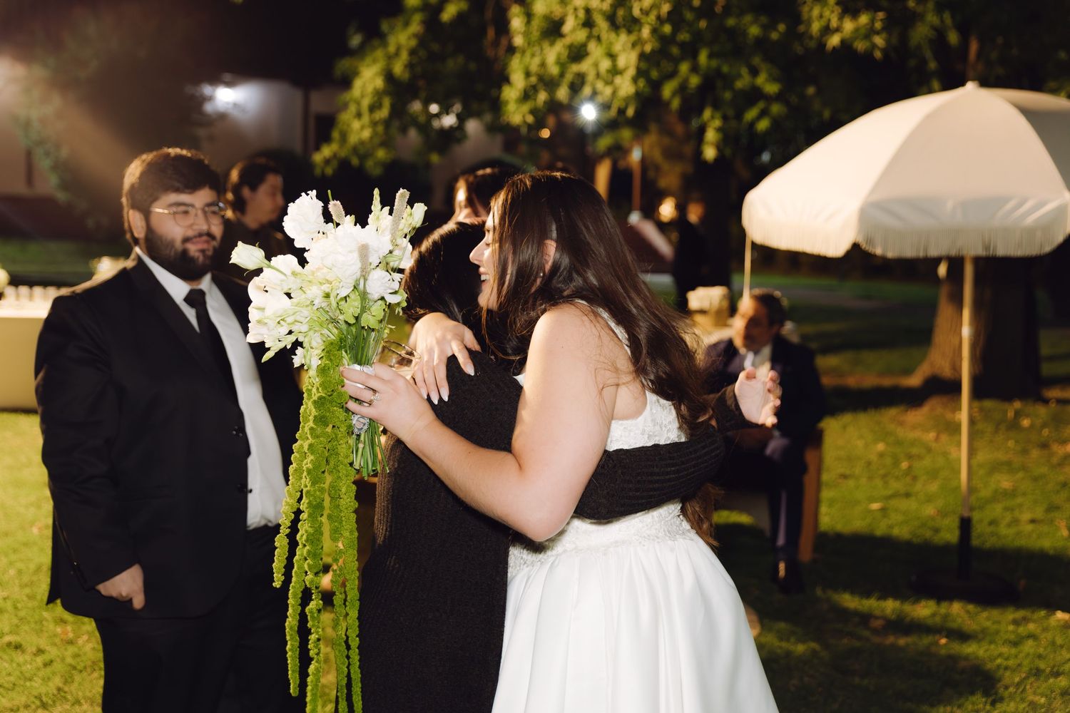 Bride and groom share a hug during outdoor evening wedding reception while guests watch nearby.