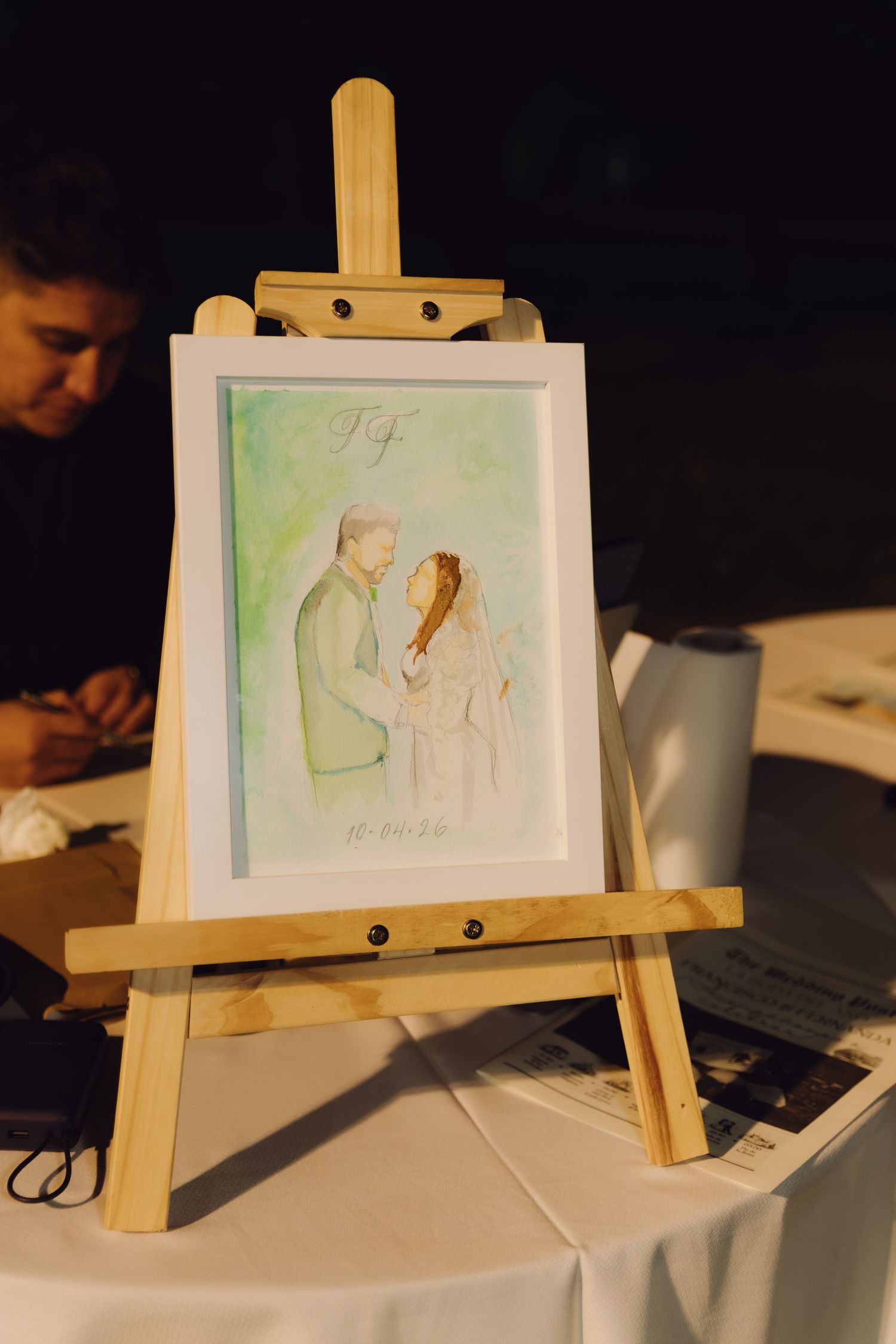 Watercolor portrait painting of bride and groom displayed on wooden easel at wedding reception.
