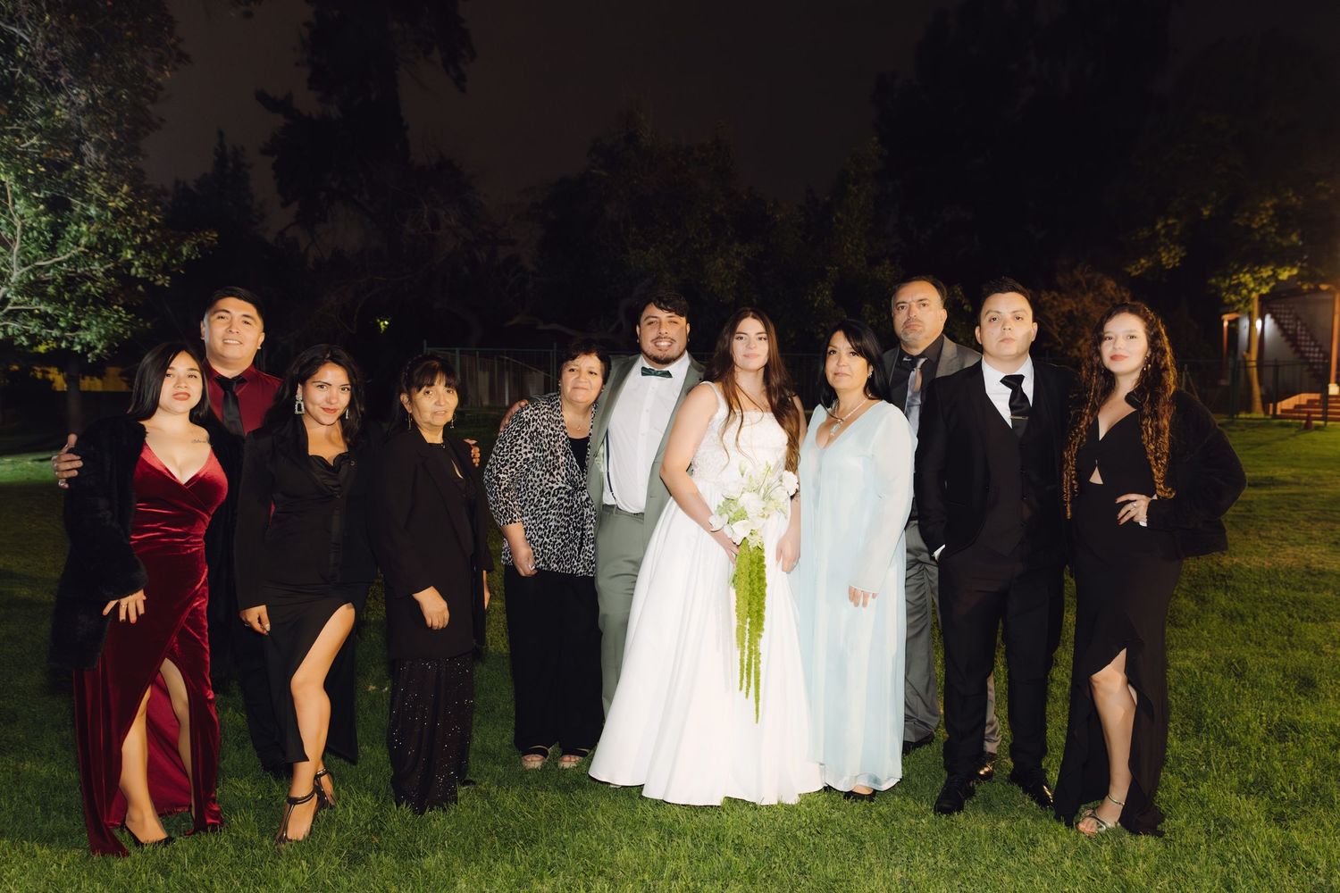 Wedding party group portrait with bride and groom posing on green lawn at night.