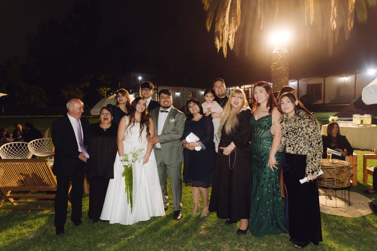 Bride and groom pose with smiling wedding guests during outdoor nighttime reception celebration.